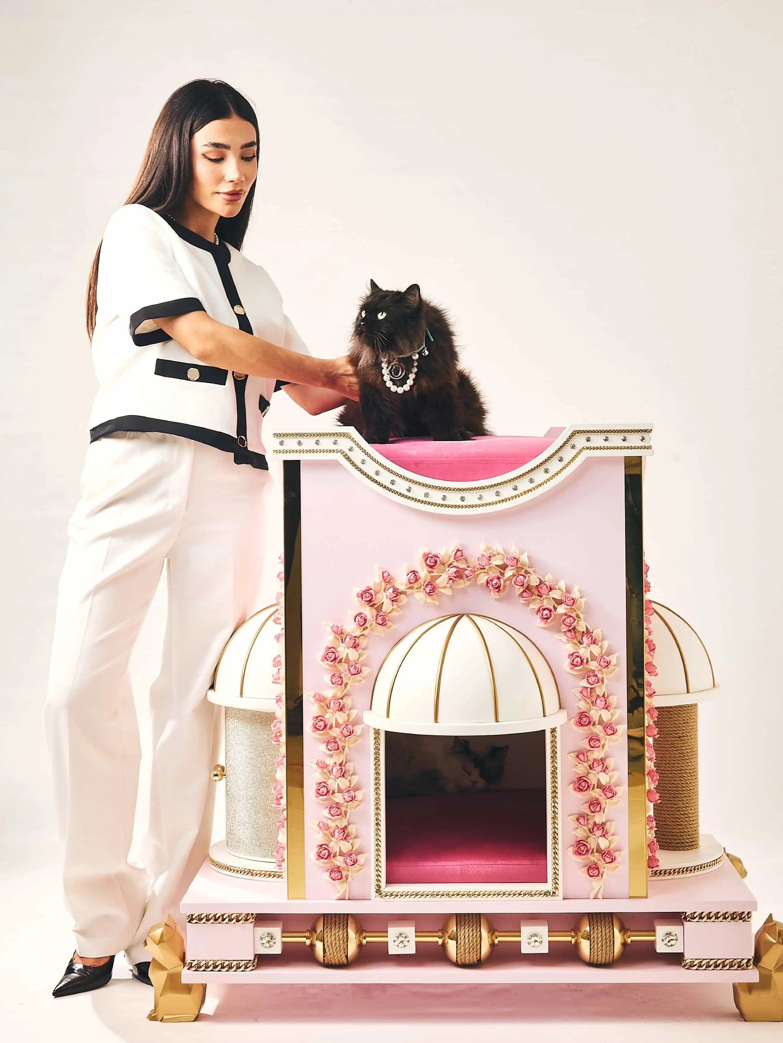 Elegant woman in white beside a luxury Furoar cat house in blush pink and gold, adorned with floral arches, as a black cat with a pearl necklace surveys its domain from the elevated perch