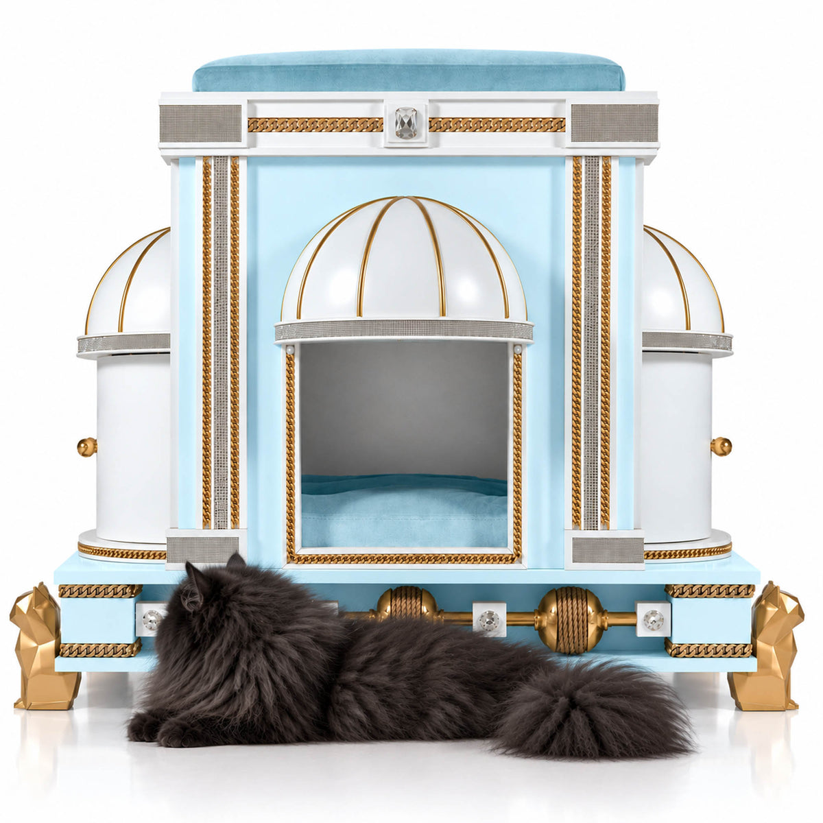 A fluffy dark gray cat lies on the floor in front of an ornate light blue and white cat bed with gold trim and dome-shaped side compartments, photographed on a clean white studio background.