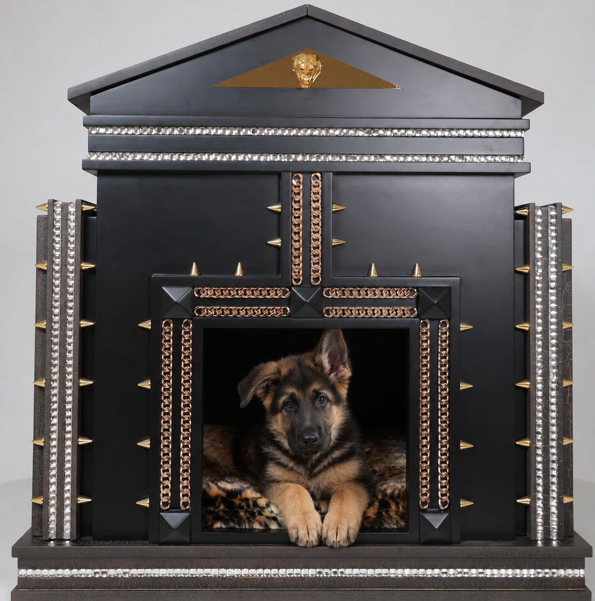 A black decorative dog house with gold and silver accents, featuring a German Shepherd lying inside on a patterned rug, photographed against a light gray studio background.