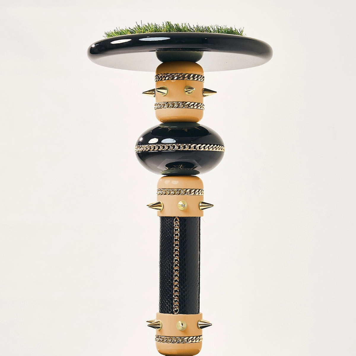 A sculptural black and gold pedestal table or candlestick-style stand with a circular top, decorative gold bands, and a small patch of green grass on the upper rim, displayed on a white pedestal against a light gray studio background.