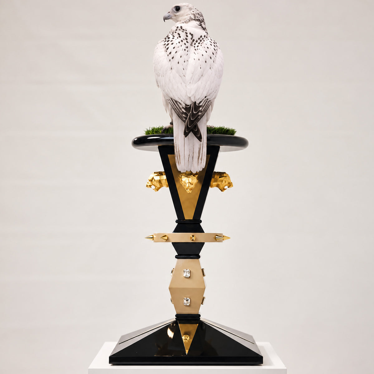 White falcon perched on the FUROAR KAISER Regal Imperial Falcon Perch, featuring a glossy black architectural base, polished gold panels, sculpted gold panther heads, crystal embellishments and a natural grass perch platform.