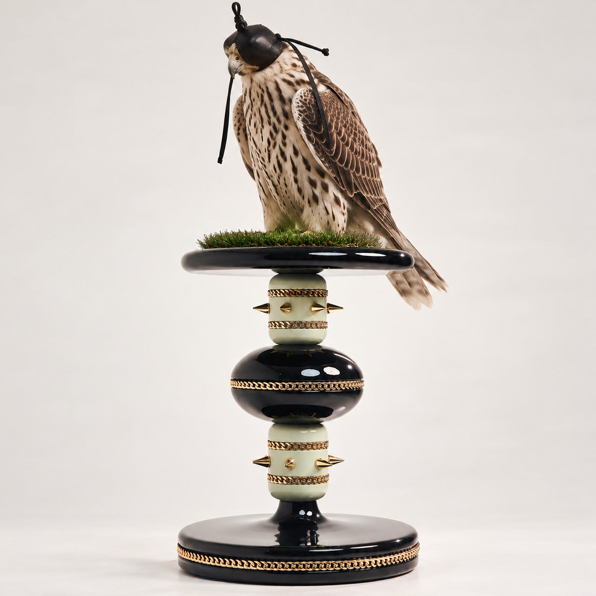Hooded falcon perched on the FUROAR SKYE Sovereign Sky Falcon Perch, featuring glossy black and ivory detailing, gold chain accents, sculptural studs and a grass-topped platform.