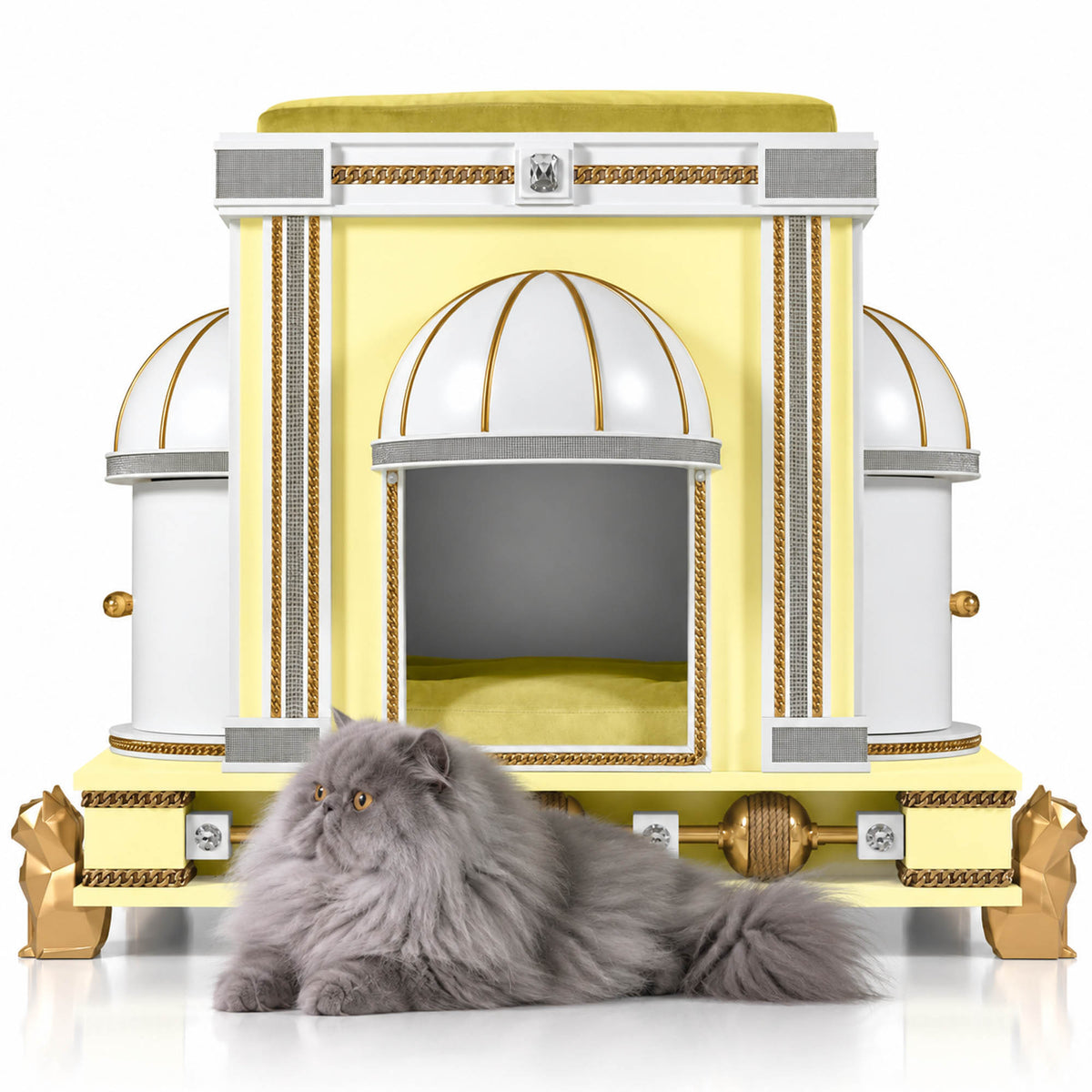 A fluffy gray long-haired cat lies in front of an ornate yellow and white cat bed designed like a classical temple, with gold trim and domed roof details, on a clean white background.