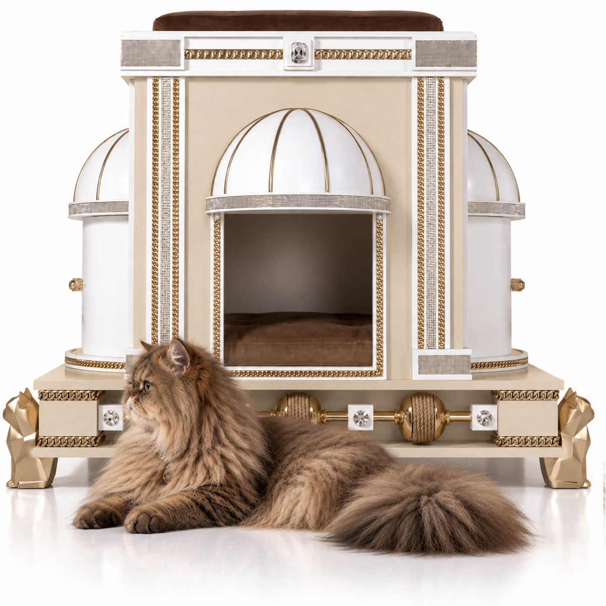 An ornate cream and gold cat house with dome-shaped openings and decorative classical detailing, shown on a white studio background with a long-haired brown cat lying in front.